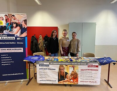 Drei gute gelaunte Mitarbeiterinnen stehen am Euro Akademie-Messestand, mit großem Roll-Up, Infobroschüren, Jutebeuteln und Stiften auf dem Tisch.