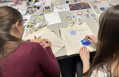 Zwei Schülerinnen sitzen an einem Tisch voller Bastelutensilien und bemalen Stofftaschen.