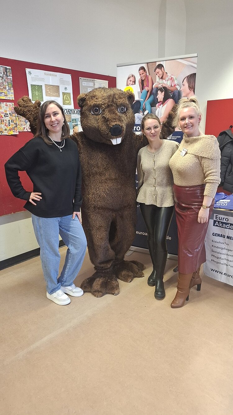 Die drei lächelnden Mitarbeiterinnen stehen am Euro Akademie-Messestand und posieren mit einem als Biber verkleideten Maskottchen für ein Bild.