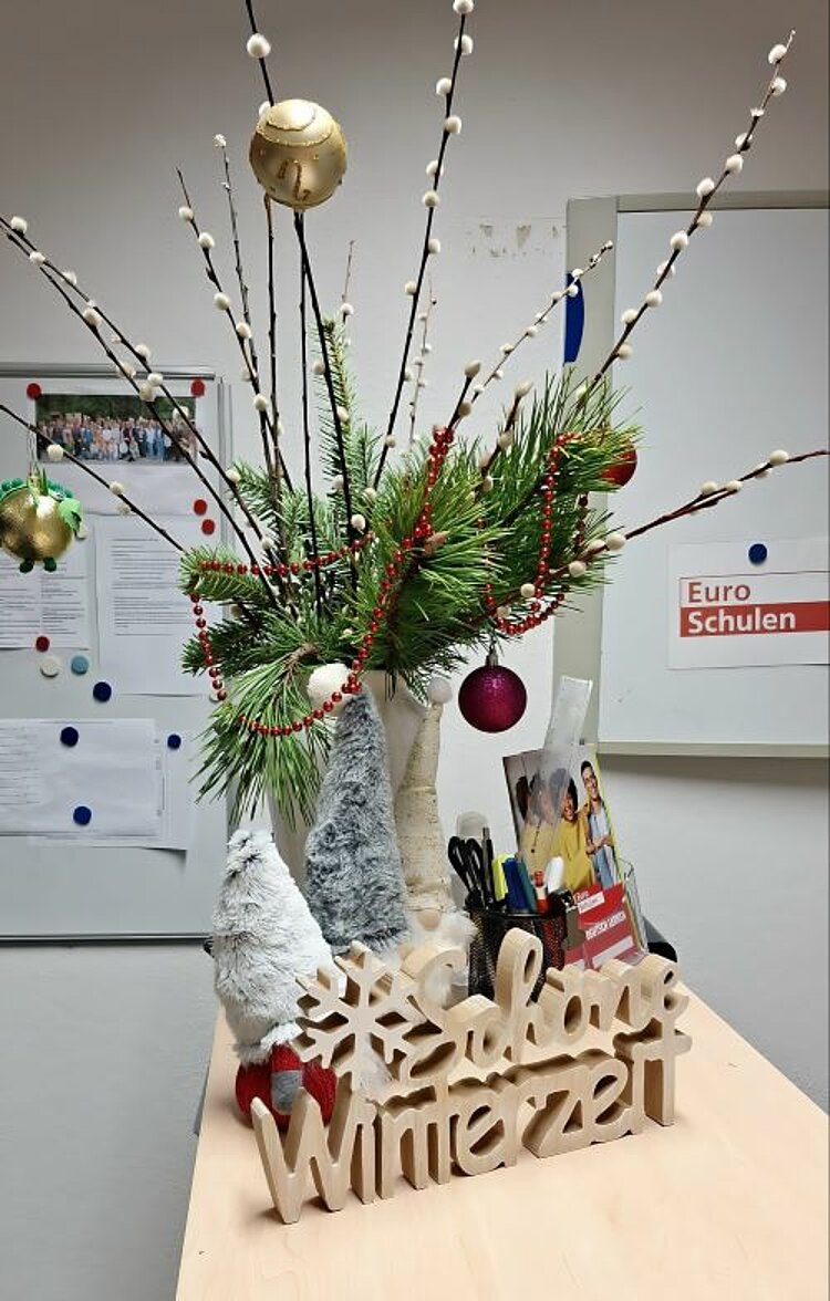 Ein weihnachtlich geschmückter Strauß aus Tannengrün und Weidenästen steht in einer weißen Vase auf einem Tisch. Davor steht eine Holzfigur, die aus den Worten "Schöne Winterzeit" und einer Schneeflocke besteht. Dahinter stehen zwei Wichtelfiguren aus Plüsch und eine Aufbewahrung mit Stiften, Scheren und Flyern von den Euro-Schulen.