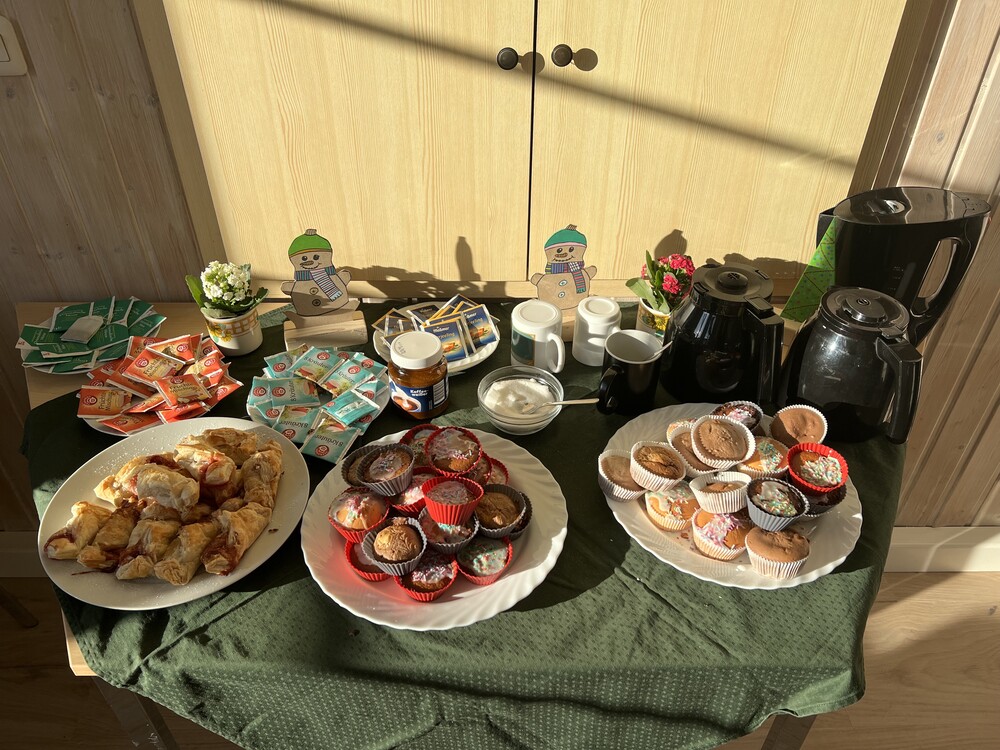 Auf einem Tisch stehen drei großes Teller mit unterschiedlichem Gebäck (u.a. Muffins) sowie mehrere Teller mit unterschiedlichen Teesorten und Zucker sowie Kannen und ein Wasserkocher. Zur Deko stehen kleine Blümchen und Schneemann-Figuren auf dem Tisch.