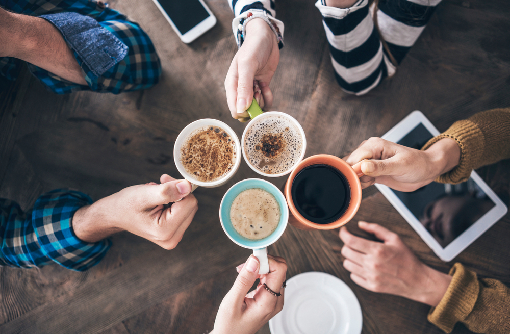 Der Blick ist von oben auf eine Holztisch gerichtet, an dem mehrere Hände mit Kaffeetassen anstoßen. Es handelt sich um Männer- und Frauenhände, die Tassen sind mit unterschiedlichen Kaffeekreation gefüllt. Außerdem liegen ein Tablet und ein Smartphone auf dem Tisch.