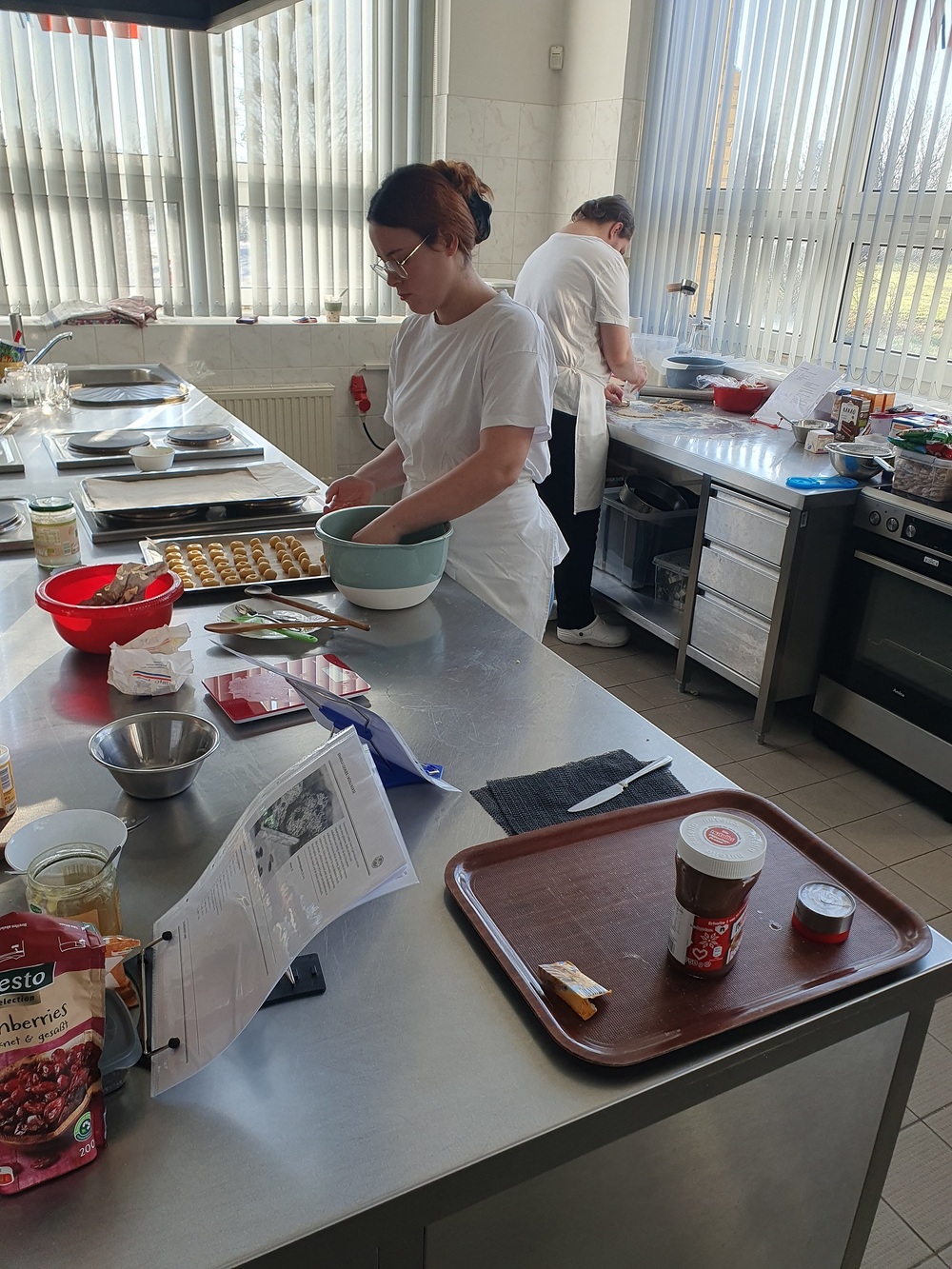 In einer Großküche arbeiten eine Schülerin und ein Schüler in weißen T-Shirts und mit Schürzen ausgestattet. Sie sind dabei unterschiedliche Plätzchen herzustellen. Auf dem Tisch stehen eine Waage, verschiedene Schüsseln und Zutaten wie z.B. Cranberries und Nussnougatcreme. Die Schülerin legt gerade kleine Kringel auf ein Backblech.