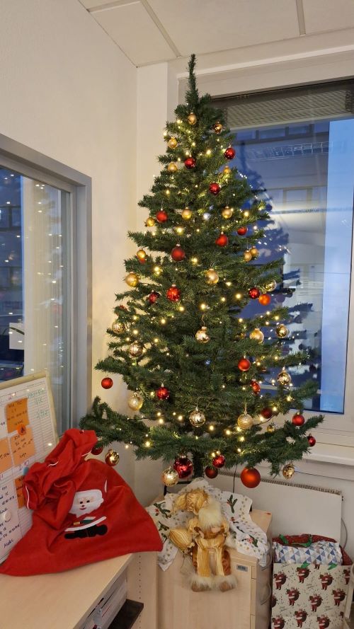 Ein Weihnachtsbaum mit roten und goldenen Kugeln sowie einer Lichterkette steht in einer Ecke eines Raums vor zwei Fenstern auf einem niedrigen Schrank. Davor sind ein Nikolausbeutel, eine Wichtelpuppe und eine Weihnachtstasche mit Geschenken drapiert.