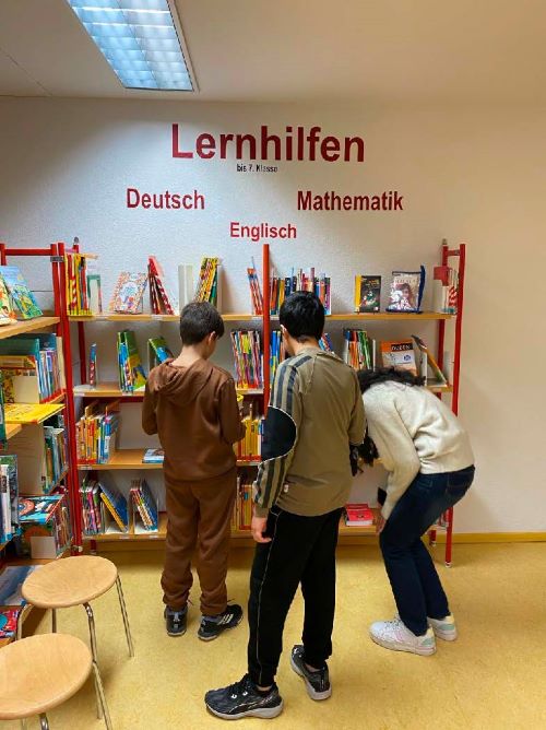 Drei Kinder stehen vor einem Bücherregal mit der Aufschrift "Lernhilfen" und den Fächern Deutsch, Englisch und Mathematik. Sie suchen sich Bücher aus.