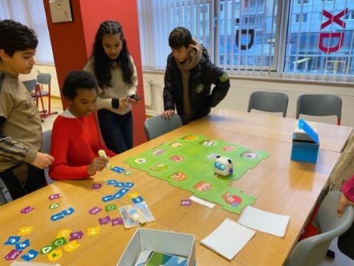 Eine Gruppe von Kindern steht um einen Tisch herum und interagiert mit einem Lernspiel, das ein Spielbrett und verschiedene Karten umfasst. Eine Roboterfigur ist auf dem Spielbrett zu sehen, und die Kinder scheinen konzentriert und engagiert zu sein.