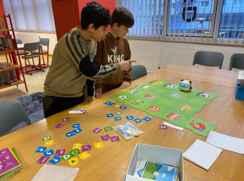 Zwei Jungen stehen an einem Tisch, auf dem ein Lernspiel mit Spielbrett und verschiedene Karten ausgebreitet ist. Eine Roboterfigur ist auf dem Spielbrett zu sehen, und die Kinder scheinen konzentriert und engagiert zu sein.