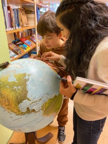 Ein Junge und ein Mädchen stehen vor einem Globus, betrachten ihn und zeigen auf einen bestimmten Punkt darauf. Im Hintergrund ist ein Regal mit Büchern zu sehen. 