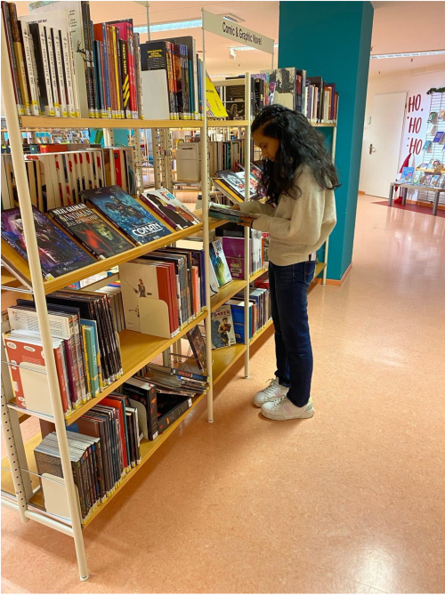 Ein Mädchen steht in einer Bibliothek und betrachtet die Bücher in einem Regal. Die Regale sind mit verschiedenen Büchern gefüllt, und im Hintergrund sind weitere Regale und ein Bereich mit Sitzgelegenheiten zu sehen.