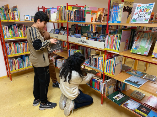 Zwei Kinder, ein Junge und ein Mädchen, stehen vor einem Bücherregal in einer Bibliothek. Der Junge hält ein Buch in der Hand und betrachtet es, während das Mädchen auf dem Boden kniet und Bücher auf den unteren Regalen durchsucht. Das Regal ist mit verschiedenen Kinderbüchern gefüllt.