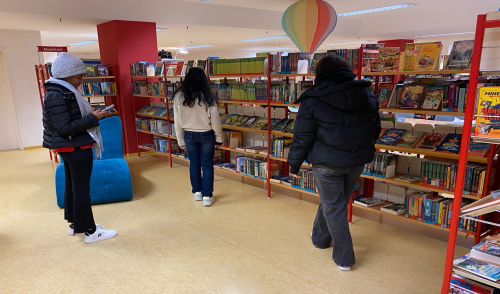 Drei Personen in einer Bibliothek, die sich Bücher in den Regalen ansehen. Eine Person steht lesend mit einem Buch im Raum, während die anderen beiden die Bücher betrachten. Im Hintergrund sind farbenfrohe Regale, ein blauer Sitzsack und ein bunter Heißluftballon an der Decke zu sehen.