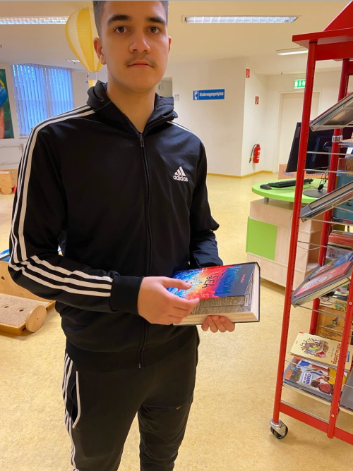 Ein junger Mann in einem schwarzen Adidas-Trainingsanzug hält ein Buch mit einem bunten Cover in einer Bibliothek..