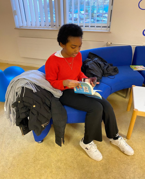 Ein Mädchen sitzt auf einem blauen Sofa und blättert in einem Buch. Neben ihr liegen eine Jacke und ein Schal.